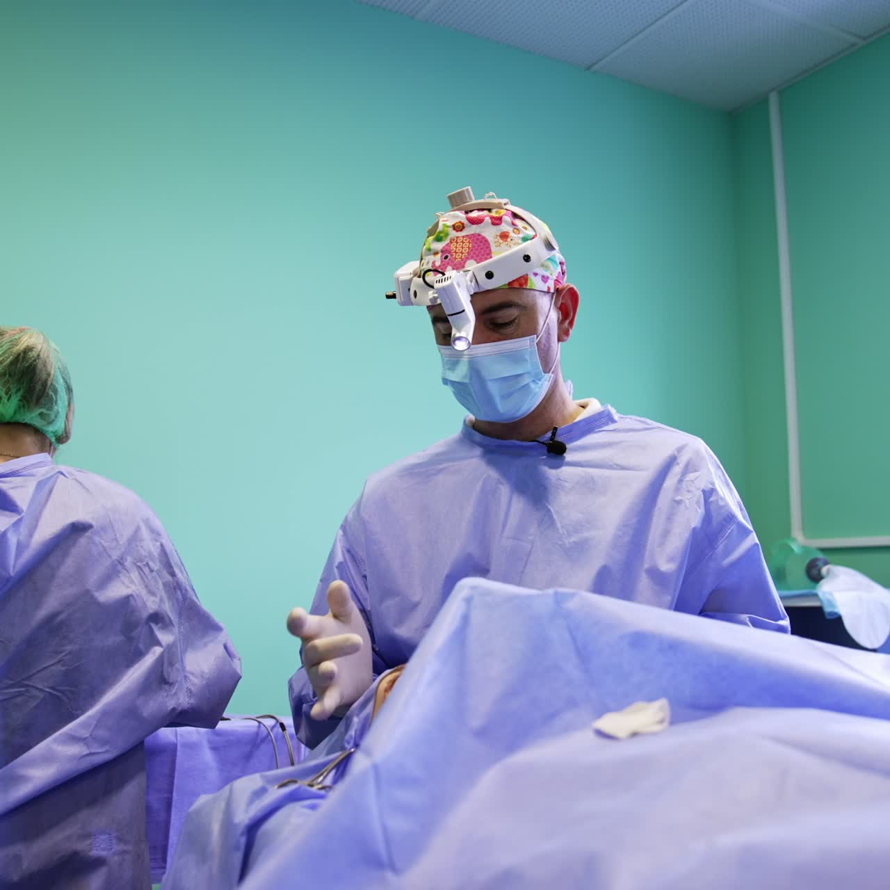 A cirurgia nas pálpebras foi finalizada. O cirurgião está perto do paciente e fala para a câmera apontando para a área operada