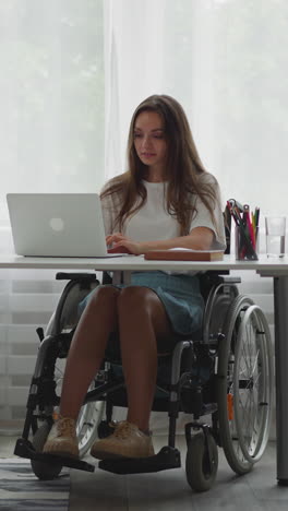 una estudiante con discapacidad se sienta en silla de ruedas haciendo la tarea en una computadora portátil moderna. una mujer con parálisis cerebral recibe educación en línea desde casa