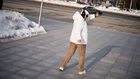 back view of woman tap dancing on busy urban street plaza in winter, white coat and boots moving rhythmically across tiled pavement, and snowy sidewalk framing dynamic outdoor performance