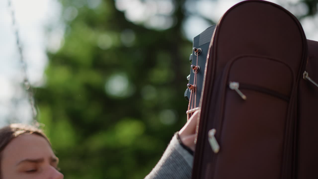 outdoor park guitarist white man adjusting guitar case, closeup of brown padded case zipper against blurred pine trees, musician face partially visible, tuning and care routine, warm sunlight, relaxed