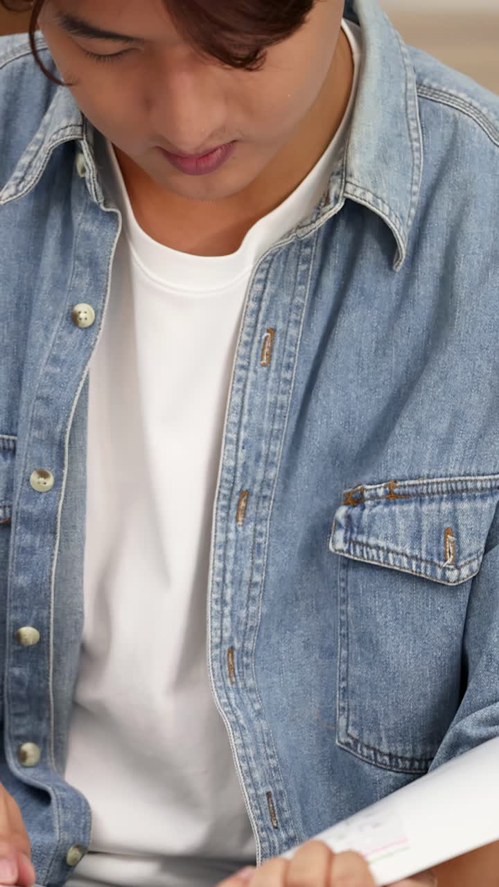 Um jovem asiático concentrado lê um livro sentado em ambiente interno, vestindo uma camisa jeans.