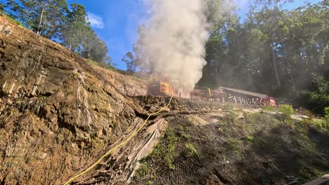 Construction vehicles and workers operate on a steep mountain road under clear skies, with visible smoke and dust