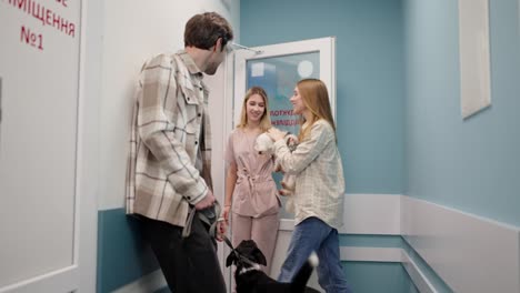 a girl veterinarian in a pink uniform invites a blonde girl with a white dog to go into the office of the veterinary clinic while the guy with his black dog remains in the corridor to wait for his turn