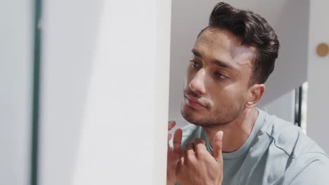 Portrait-of-happy-biracial-man-examining-face-and-skin-in-bathroom-mirror-and-smiling,-slow-motion