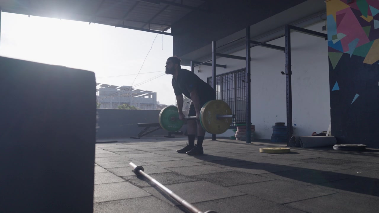 Brodaty sportowiec płci męskiej z Azji Południowej wykonujący konwencjonalny martwy ciąg ze sztangą podczas treningu siłowego w nowoczesnym, krytym studiu fitness. Kolekcja: WORKOUTSERIES2026