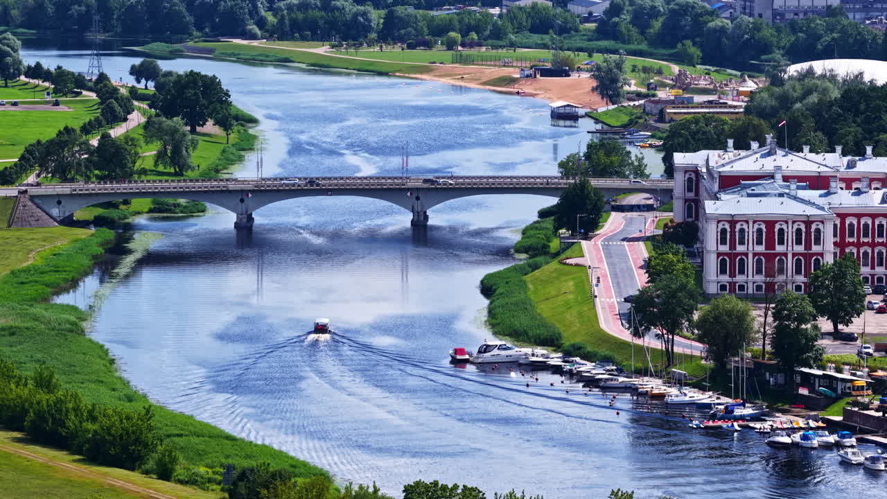 Aerial view of Jelgava Palace beside the Lielupe River, with boats moving across the water and a historic landmark surrounded by green parks and bridges. A scenic Latvian city landscape