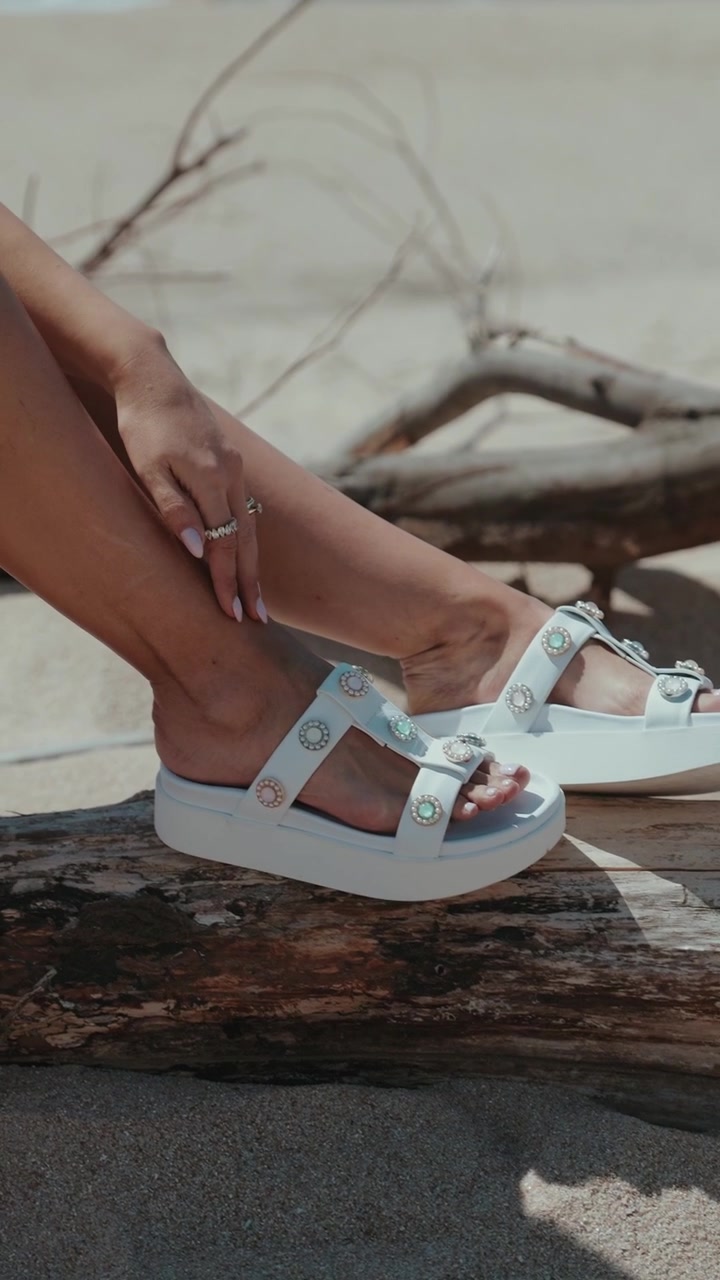 Gambe femminili con sandali bianchi appoggiate su legno galleggiante su una spiaggia sabbiosa.