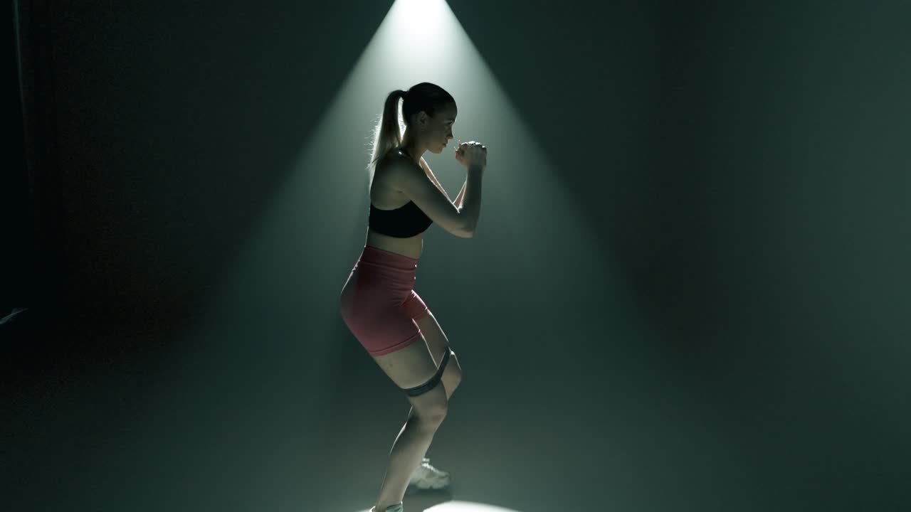 Blonde woman in gym performs side lunges with resistance band above knees. Cinematic light creates glowing rim on her body. Full body tracking shot from feet to head in smoky dark training studio