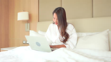 in a work from anywhere world, a woman in a bathrobe sits up in bed while working on her laptop