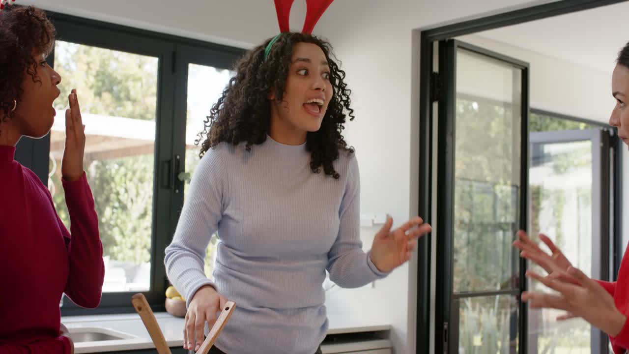 Diverse female friends reacting to red sweater gesture, friend picking spoon whispering in kitchen