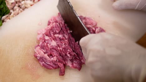 hand with glove use knife to slice beef into pieces on white chopping board, close up