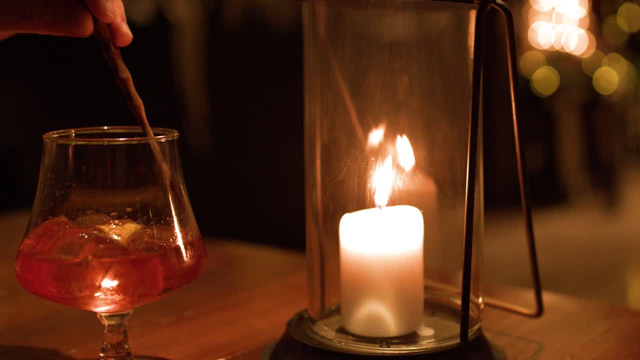 Una mano remueve un cóctel rojo en un vaso junto a un farol con una vela encendida.