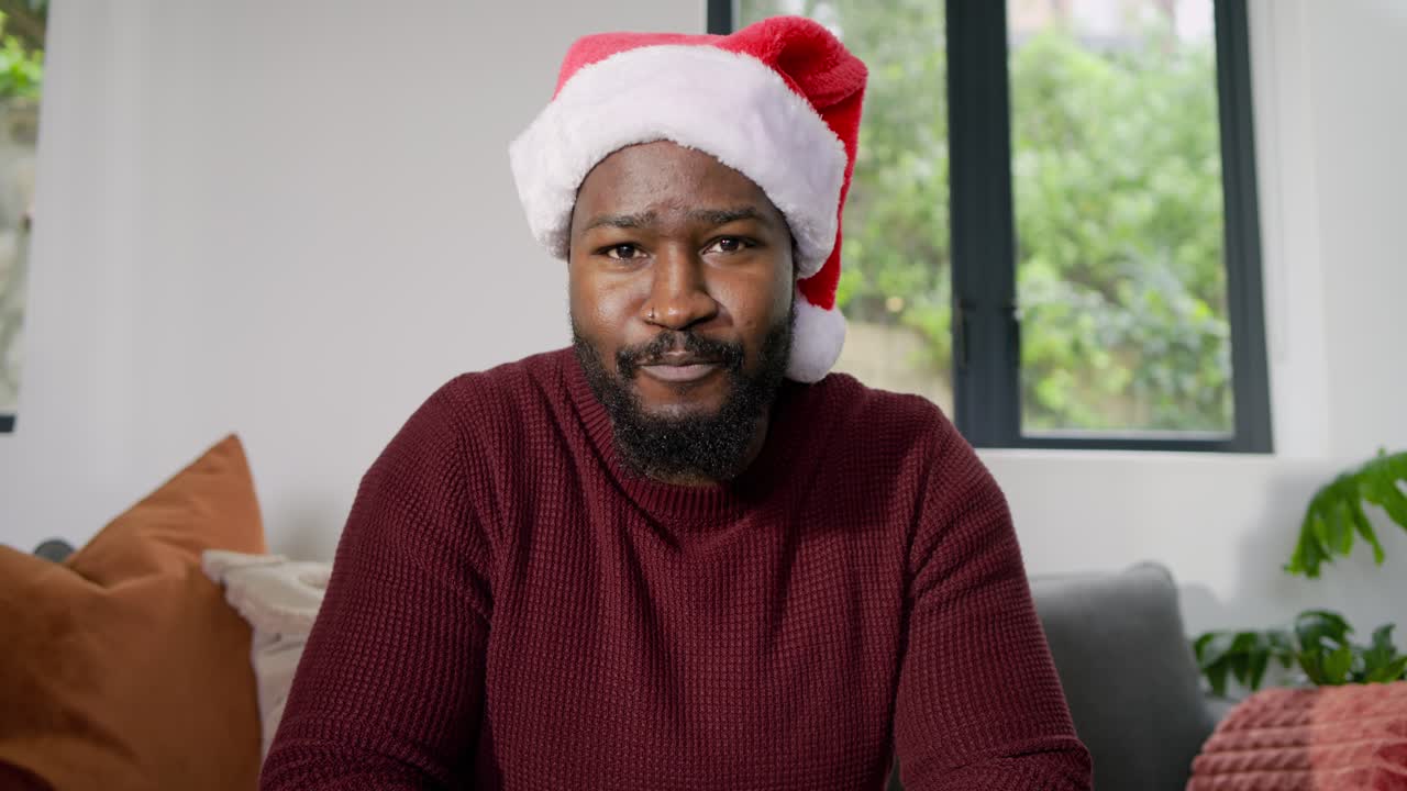 Lifting gift African American man wearing Santa hat speaking holiday greetings on gray sofa