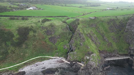 Dolly-backwards-aerial-drone-shot-of-Antrim-County-Northern-Ireland-Gobbins-overlooking-a-rocky-path,-the-beautiful-green-landscape-and-a-gorgeous-blue-river-in-the-morning