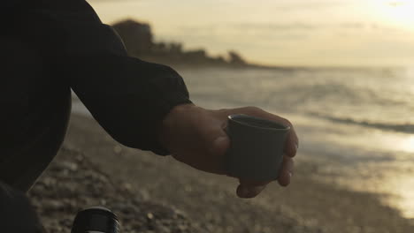 Man-pours-coffee-from-thermos-at-beach-with-sea-waves-golden-bay-blurry-background