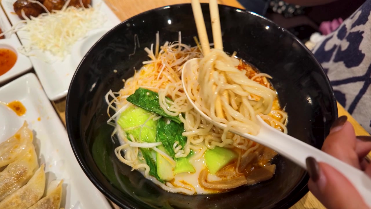 Close-up tangan menggunakan sumpit dan sendok untuk makan mi ramen dalam kuah pedas.