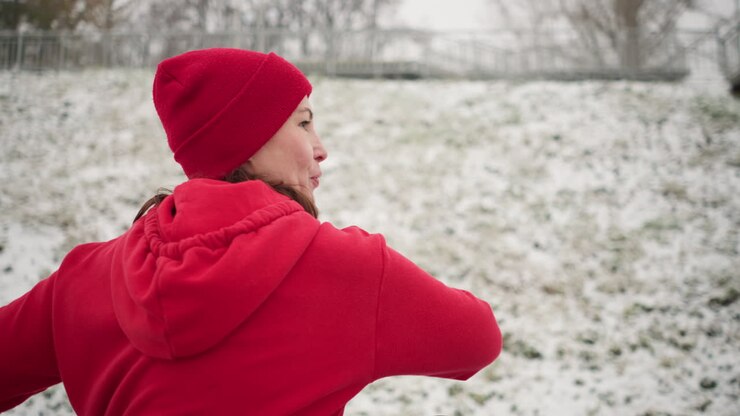 Woman performs outdoor workout with hand up for balance, turning left and right for coordination and strength, wearing red hoodie and black gloves, surrounded by snowy ground