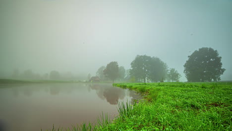 Zeitrafferaufnahme-Des-Morgennebels,-Der-Morgens-über-Dem-Von-Grüner-Vegetation-Umgebenen-See-Aufsteigt