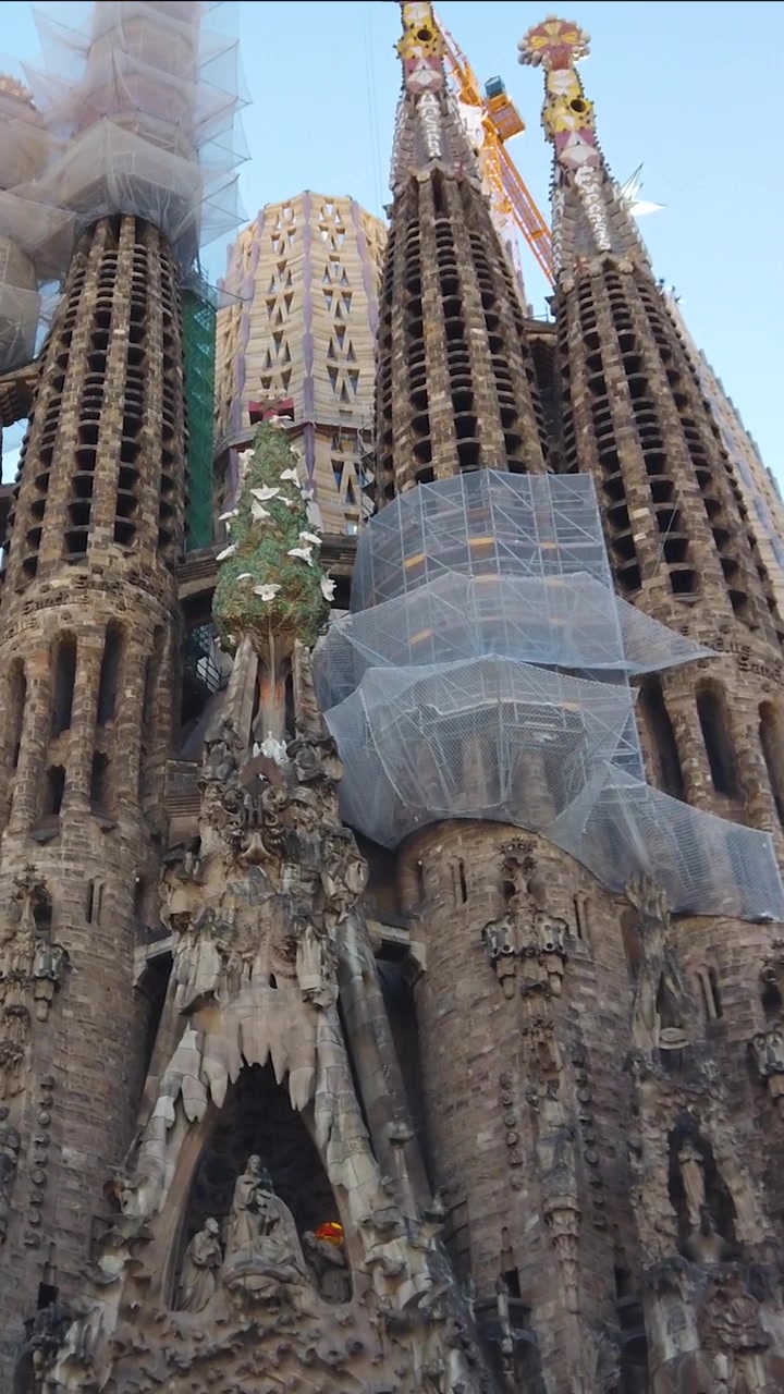 Panoramica verso il basso sulla Sagrada Familia che rivela guglie ornate, impalcature e intricate facciate in pietra ancora in costruzione, catturando l'ambizione secolare del capolavoro di Gaudí a Barcellona