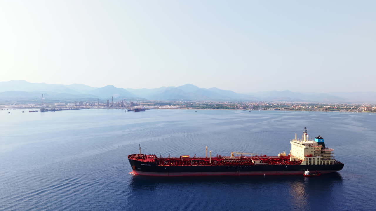Drone advances toward oil tanker offshore Sicily with refinery, mountains, and moving boat in the distance