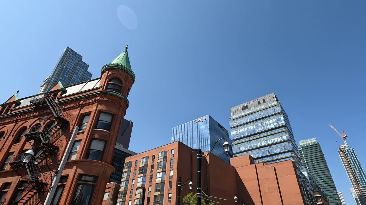 Moving shot capturing the historic Flatiron Building contrasted with modern skyscrapers in Toronto, Canada, under a bright, cloudless sky. A unique blend of old and new architecture