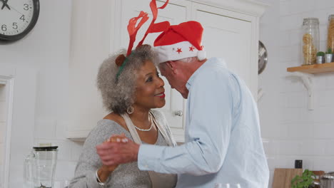 Loving-Senior-Couple-Wearing-Fancy-Dress-Antlers-Dance-In-Kitchen-Whilst-Preparing-Christmas-Dinner