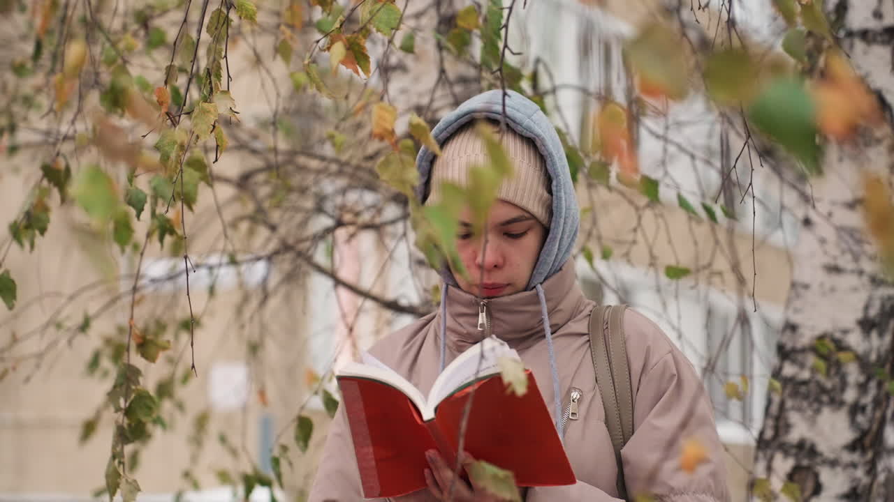 Light skin lady wearing beige winter jacket and beanie stands under tree with autumn leaves, attentively flipping through red book pages in calm outdoor setting with soft natural light