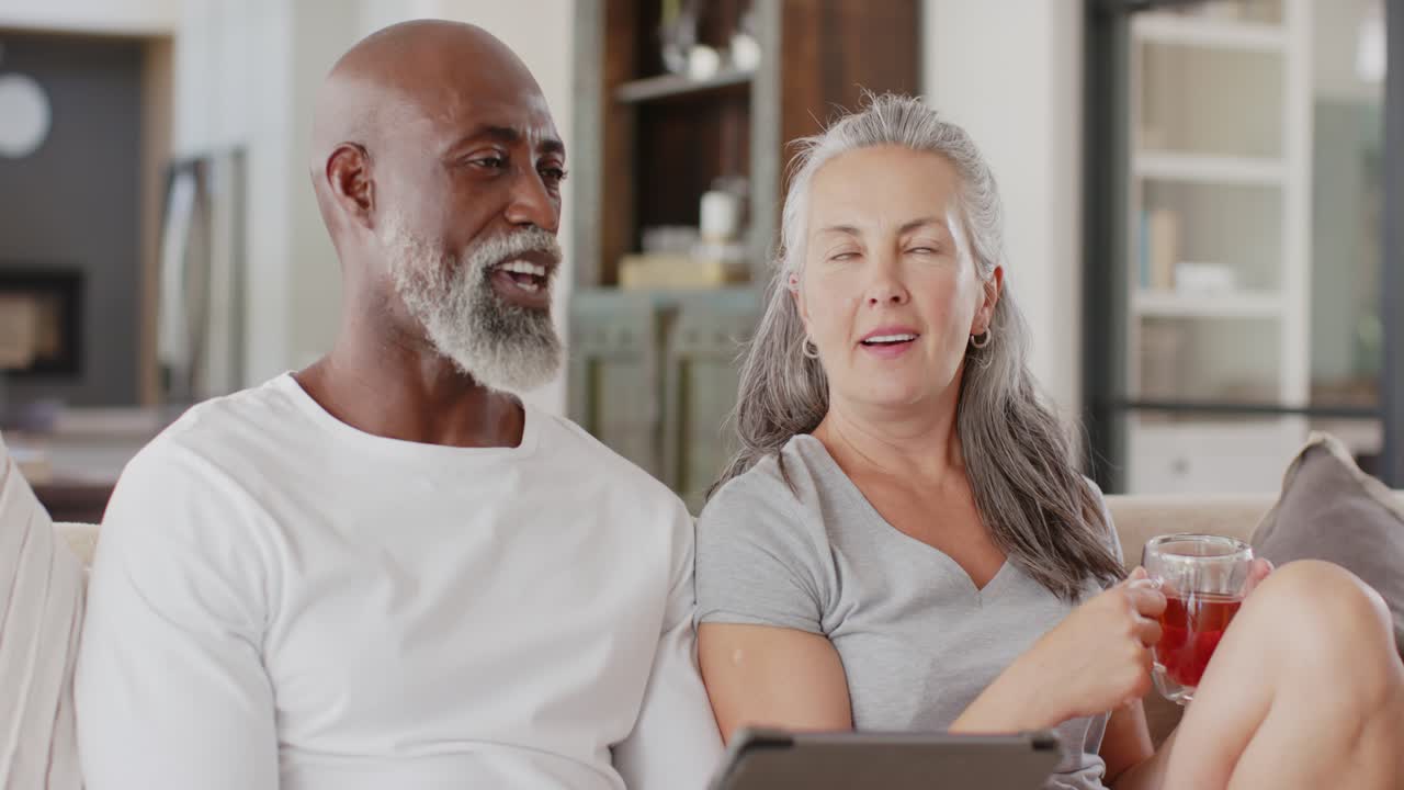 Senior couple sitting on couch holding mug and tablet, reacting to off-camera noise, laughing