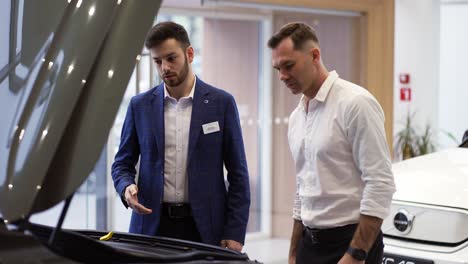 consultant showing car engine under open hood to a client in auto showroom