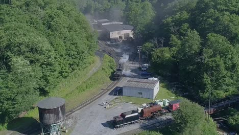 an aerial view of an antique steam locomotive backing up to a water tower to take a drink on a sunny summer morning