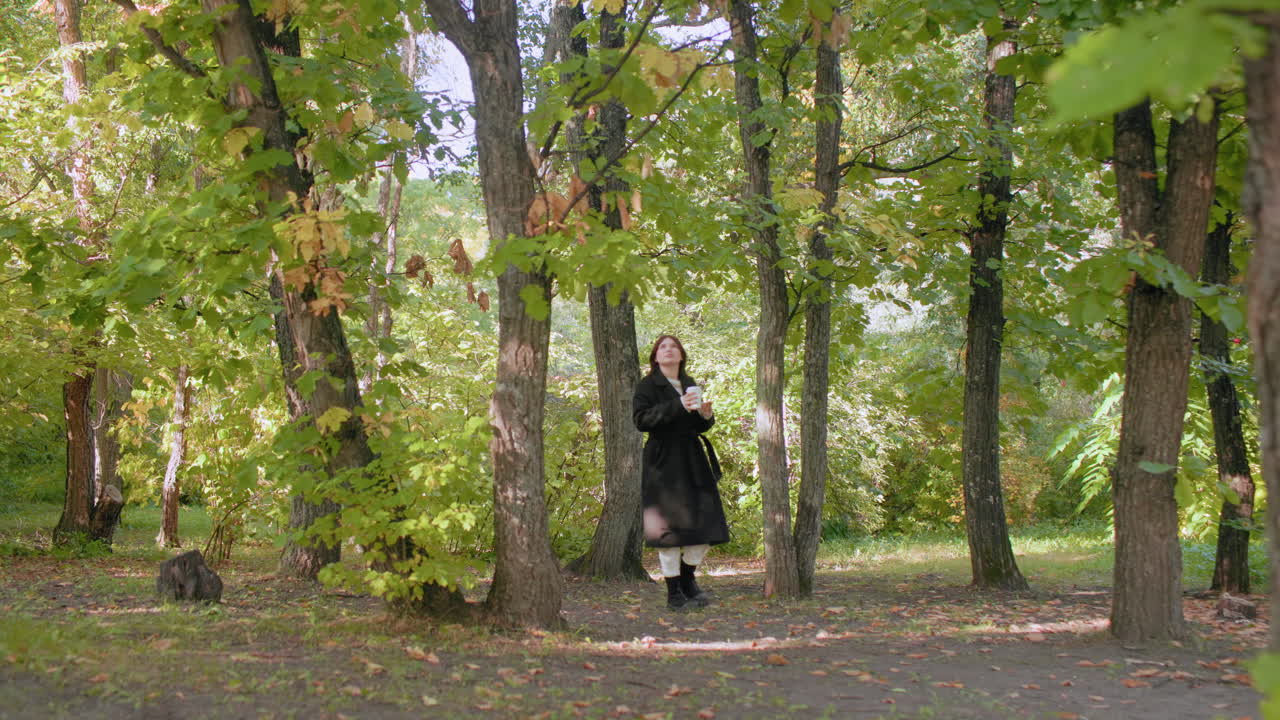 Широкий вид на гуляющего по парку человека, попивающего горячий чай из кружки во время прогулки под лиственным навесом, остановившегося среди зеленых деревьев и пятнистого солнечного света, медленное расслабленное движение, запечатленное в безмятежной лесной обстановке.