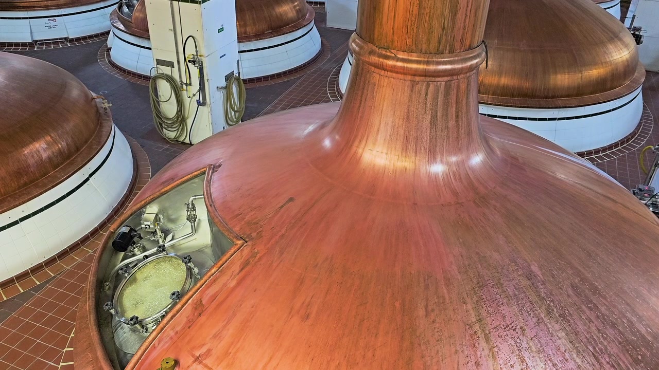 Close view of large copper beer vats in a brewery, showing fermentation equipment, tiled production room, round brew kettles, metal fittings, hoses, and polished industrial details.