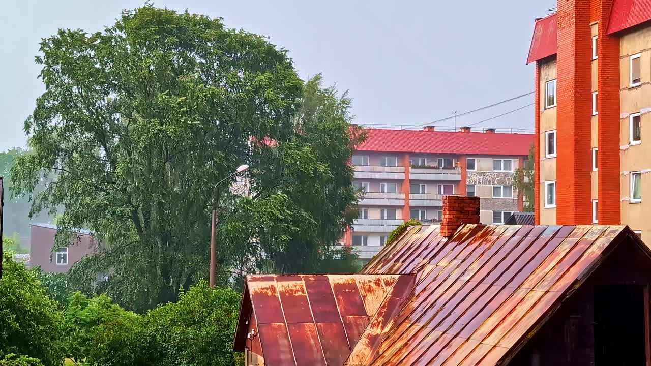 Rain falling on rusty metal roof with urban buildings and lush green trees
