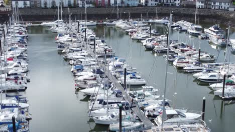 Scenic-luxurious-waterfront-harbour-apartment-village-yachts-and-sailboats-under-mountain-coastline-aerial-lowering-right-tilt-up-view