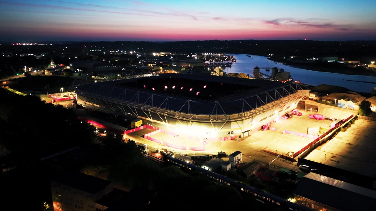 Nært droneflyfoto av den sterkt opplyste forplassen og taket til St Marys Stadium med Southampton bylys og elven Itchen i bakgrunnen.