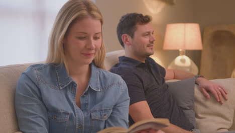 evening shot of couple relaxing on sofa at home with woman reading book and man watching tv - shot in slow motion