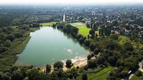 Explore-Un-Paisaje-Veraniego-Bañado-Por-El-Sol-Con-Un-Dron:-Un-Extenso-Campo,-Un-Lago-Sereno,-Bañistas,-Una-Animada-Cancha-De-Baloncesto-Y-Un-Animado-Campo-De-Fútbol.