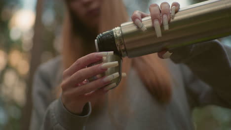 close-up of young woman pouring water from thermos into its cap in peaceful forest setting, capturing her well-manicured nails drinks from it amidst blurred greenery background