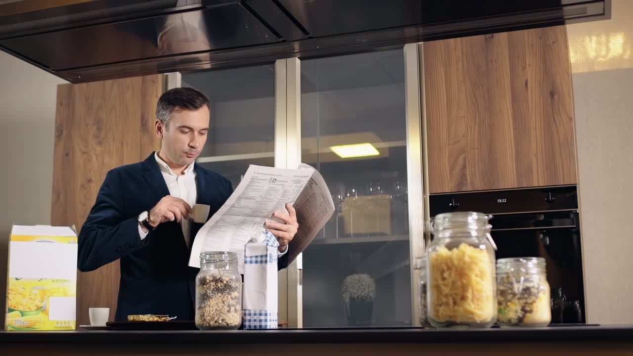 A caucasian man in jacket drinking coffee and reading a newspaper on a kitchen. Table with food