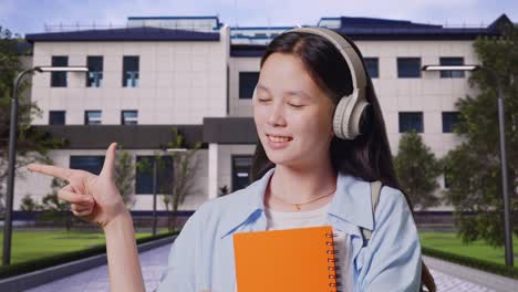 close up de una estudiante adolescente asiática con una mochila y algunos libros sonriendo y señalando a un lado mientras está de pie frente a un edificio escolar