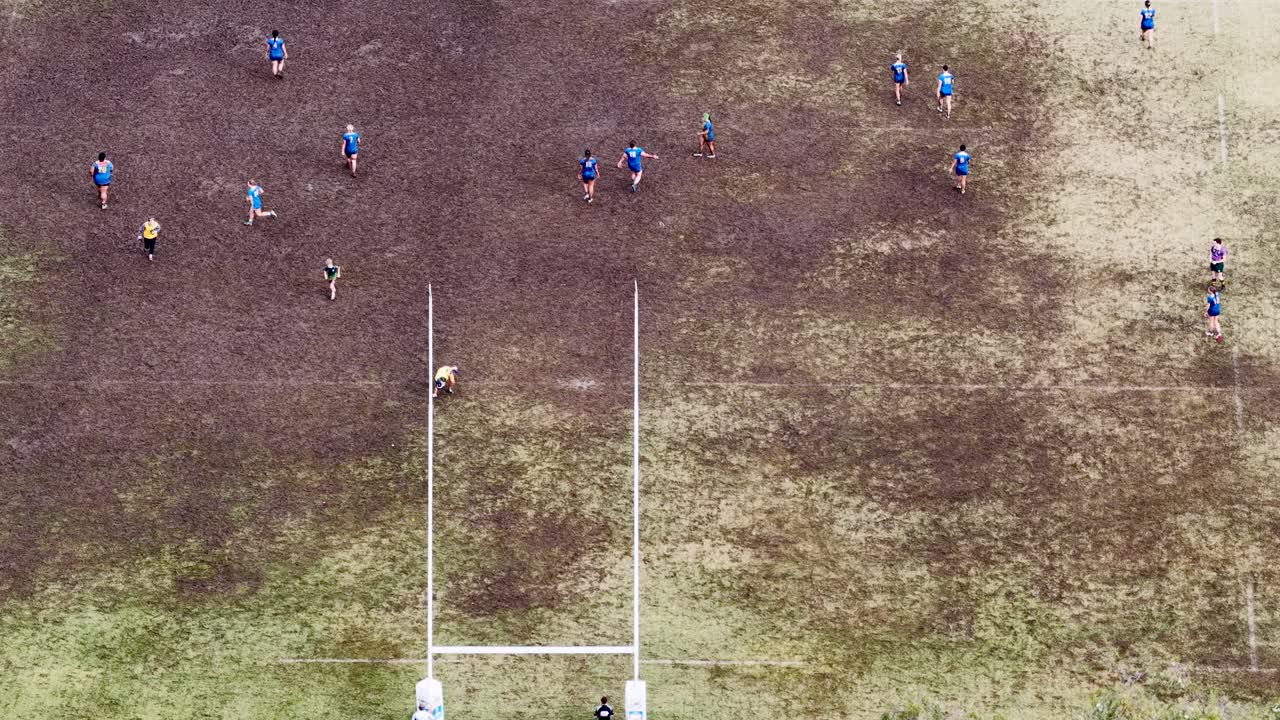Children engage in a lively rugby match on a muddy field, captured from above. Overcast lighting and dynamic movement create an energetic atmosphere