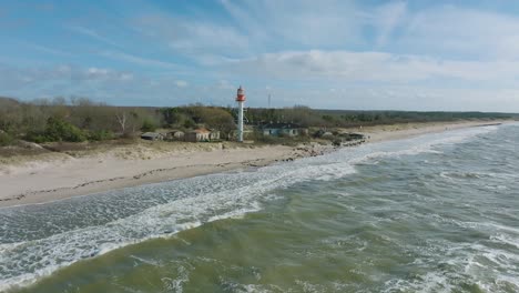 Aerial-establishing-view-of-white-colored-Pape-lighthouse,-Baltic-sea-coastline,-Latvia,-white-sand-beach,-large-waves-crashing,-sunny-day-with-clouds,-wide-drone-shot-moving-forward,-camera-tilt-down