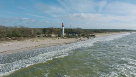 aerial establishing view of white colored pape lighthouse, baltic sea coastline, latvia, white sand beach, large waves crashing, sunny day with clouds, wide drone shot moving forward, camera tilt down