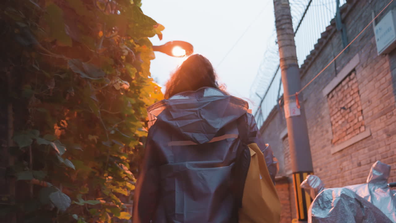 Mujer asiática caminando por un callejón, repartidor encapuchado navegando un estrecho pasaje de ladrillo, pasando una caja de entrega roja y un patinete aparcado, deteniéndose bajo el cálido resplandor de una lámpara, paredes texturizadas y enredaderas crean un ambiente íntimo