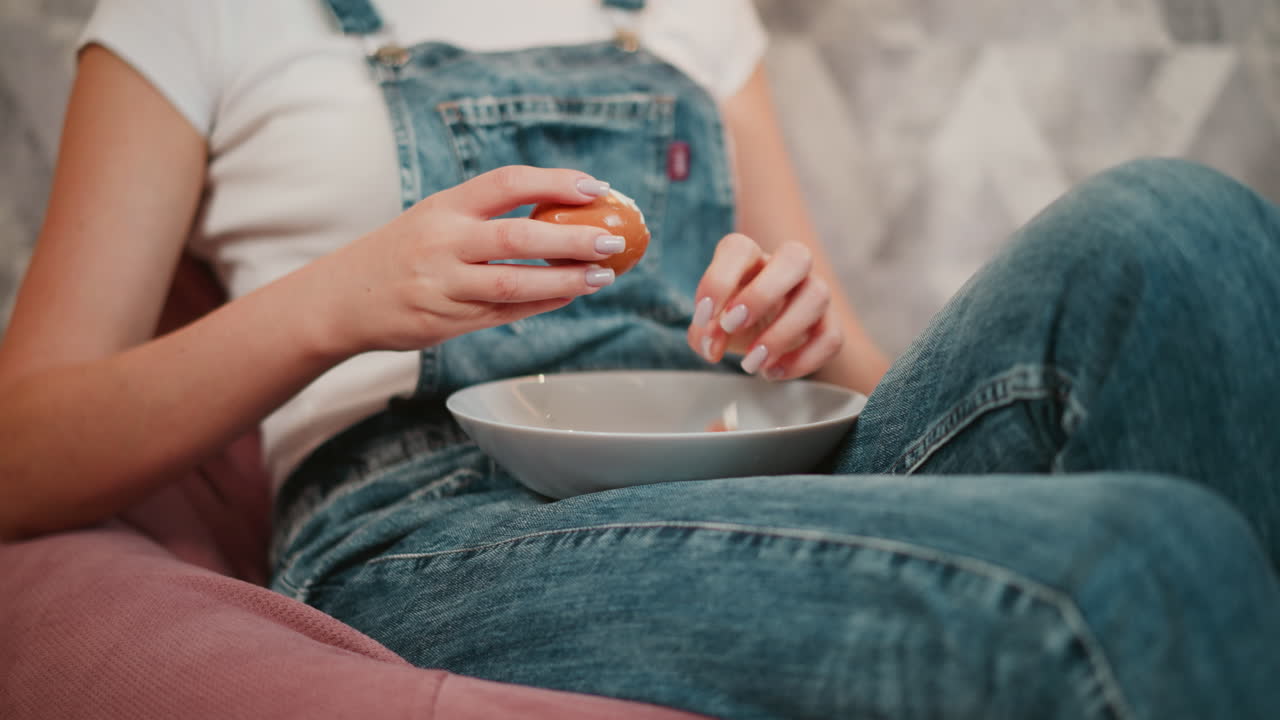 Close up of woman in denim overalls sitting with legs crossed, gently peeling brown boiled egg over glass plate resting on her lap, captured in cozy indoor environment with soft lighting