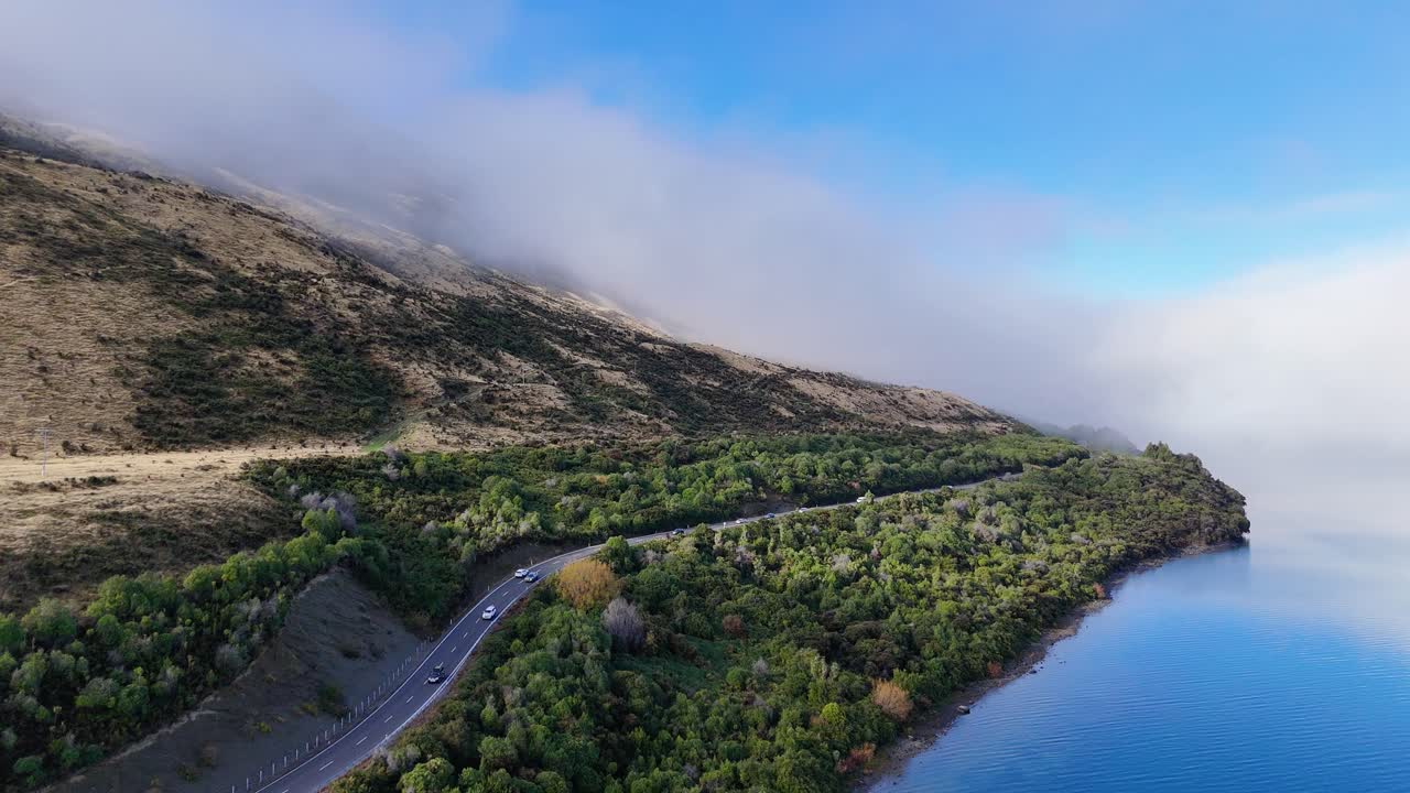 Aerial view of a winding road beside Lake Wakatipu, surrounded by lush greenery and mountains under a clear blue sky