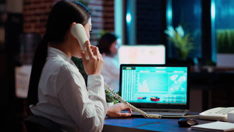 Employee-taking-landline-telephone-call-while-working-with-financial-graphs