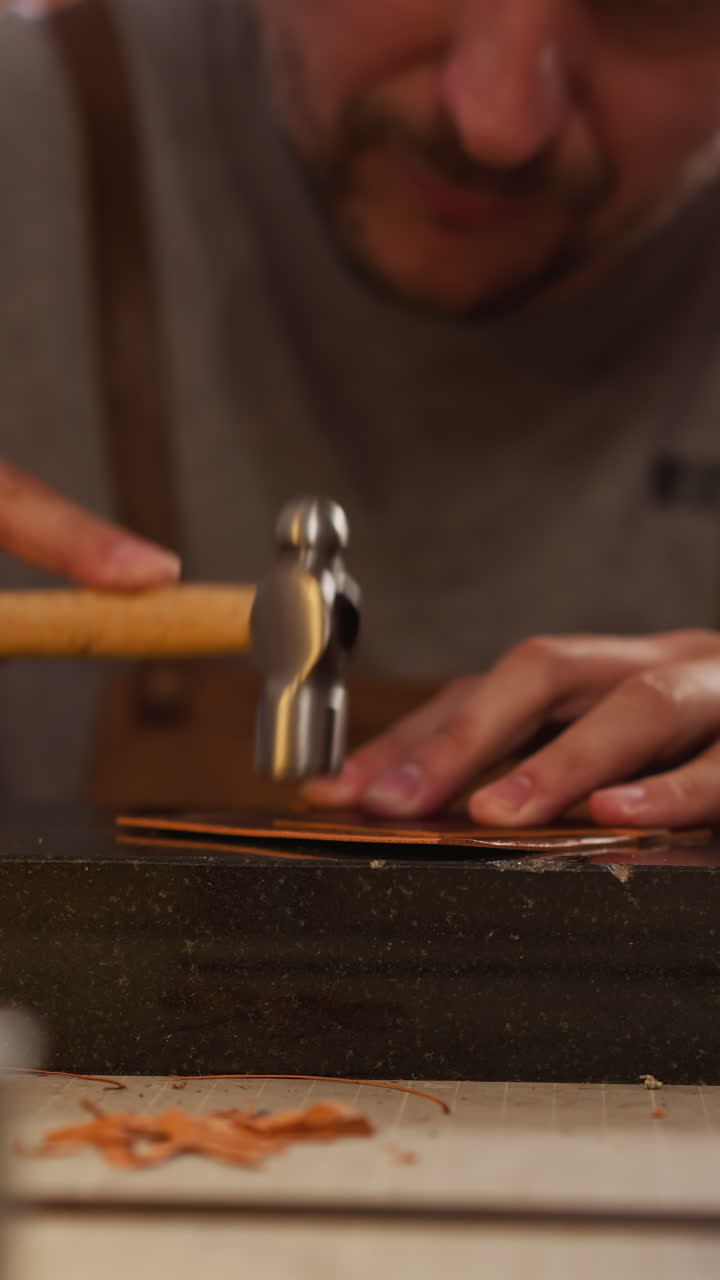 Concentrated leathersmith processes sheet of material with hammer at table in workshop closeup. Making elegant accessories from natural leather