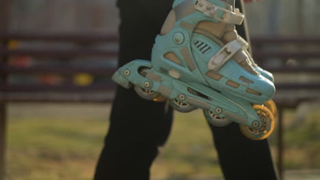 close-up of a hand holding a pair of rollerblades while walking through a blurred park. the person is wearing black trousers, and the focus is on the rollerblades against a soft, blur background