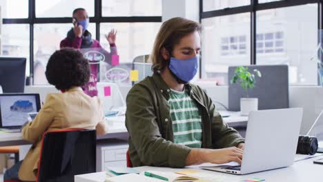 caucasian businessman wearing face mask sitting using a laptop in modern office